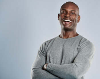 Laughing single handsome young African man with bald head in gray long sleeve shirt and folded arms over neutral background